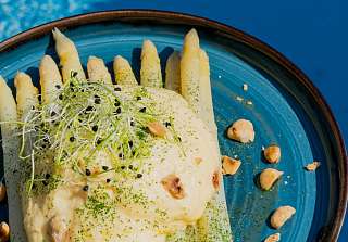 Weisser Spargel mit Sprossen, Kräuteröl und gerösteten Nüssen, serviert auf einem blauen Teller