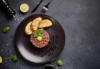 Rindfleisch-Tatar mit Basilikum, Kapern und geröstetem Weissbrot, serviert auf einem dunklen Teller