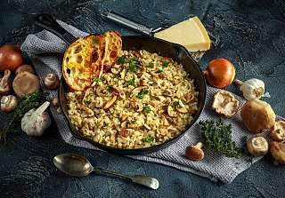 Steinpilzrisotto mit frischen Kräutern in einer Eisenpfanne, umgeben von Steinpilzen, Zwiebeln, Knoblauch, Thymian und Parmesan